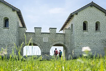 Zwei Personen mit Rucksäcken stehen unter einem steinernen Torbogen einer historischen Anlage bei sonnigem Himmel.