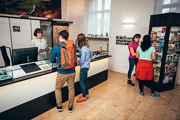 Innenraum einer Touristeninformation mit vier Personen, zwei am Tresen und zwei vor einem Prospektständer.