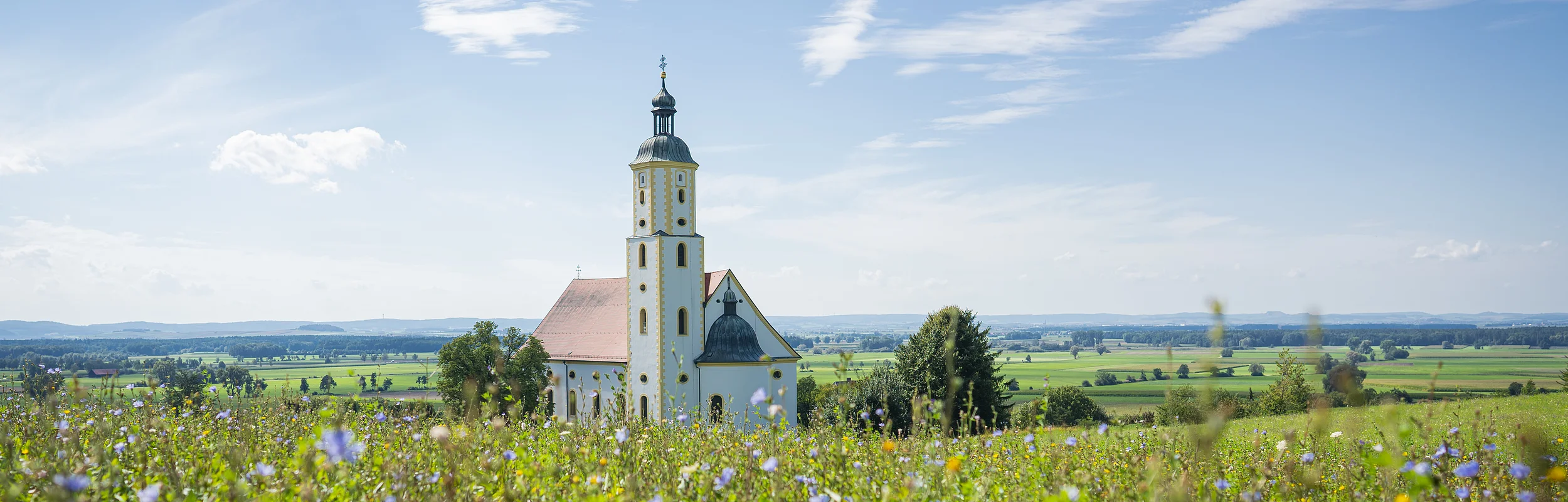 Kirche mit Turm hinter blühendem Feld unter blauem Himmel mit Wolken in ländlicher Umgebung