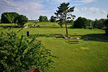 Grüne Wiesen mit Baum, Ruinenfundamenten und blauem Himmel mit Wolken im Hintergrund.