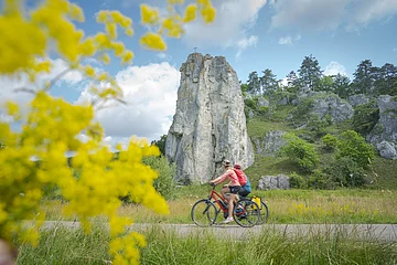 Zwei Radfahrer fahren auf einem Weg vor einem hohen Felsen mit Kreuz, gelbe Blumen im Vordergrund, bewölkter Himmel