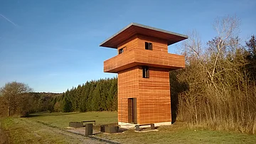 Holzbeobachtungsturm mit umlaufendem Balkon in einer Wiesenlandschaft neben Wald und kahlen Bäumen bei blauem Himmel
