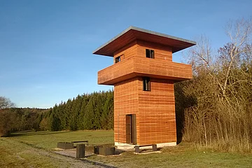 Holzbeobachtungsturm mit umlaufendem Balkon in einer Wiesenlandschaft neben Wald und kahlen Bäumen bei blauem Himmel