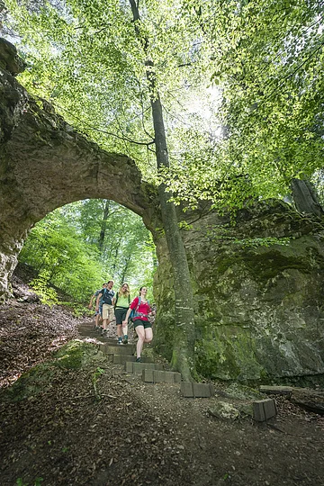 Vier Wanderer gehen einen Waldweg mit Steintreppe unter einem natürlichen Felsbogen hinab.