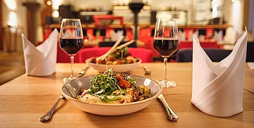 Tisch mit zwei Tellern, zwei Gläsern Rotwein, Besteck und zwei gefalteten weißen Servietten in einem Restaurant.