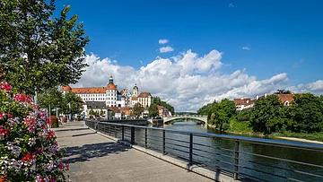 Fluss mit Brücke, rechts Bäume und Häuser, links Blumen, Baum und Schloss im Hintergrund bei blauem Himmel.