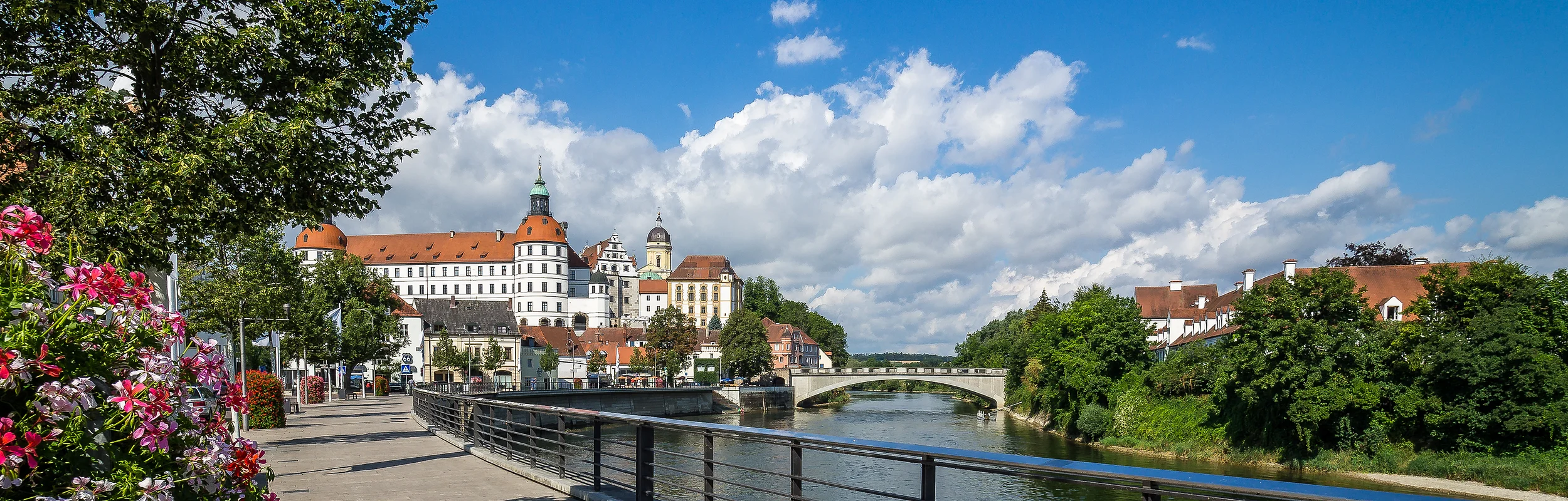 Fluss mit Brücke, rechts Bäume und Häuser, links Blumen, Baum und Schloss im Hintergrund bei blauem Himmel.