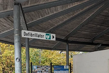 Überdachter Rastplatz mit Holzbänken, Infotafeln und einem Defibrillator an der Wand.