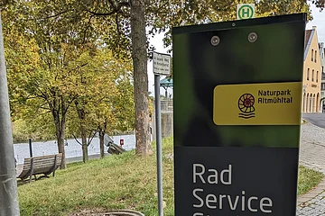 Fahrradservicestation mit Werkzeug- und Fahrradsymbol, im Hintergrund Bäume und ein Fluss.