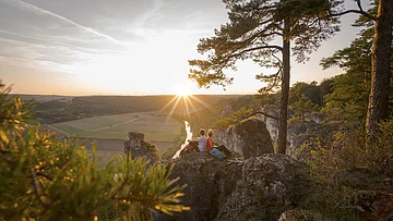 Zwei Personen sitzen auf Felsen und schauen bei Sonnenuntergang auf ein Flusstal mit Bäumen und Feldern.
