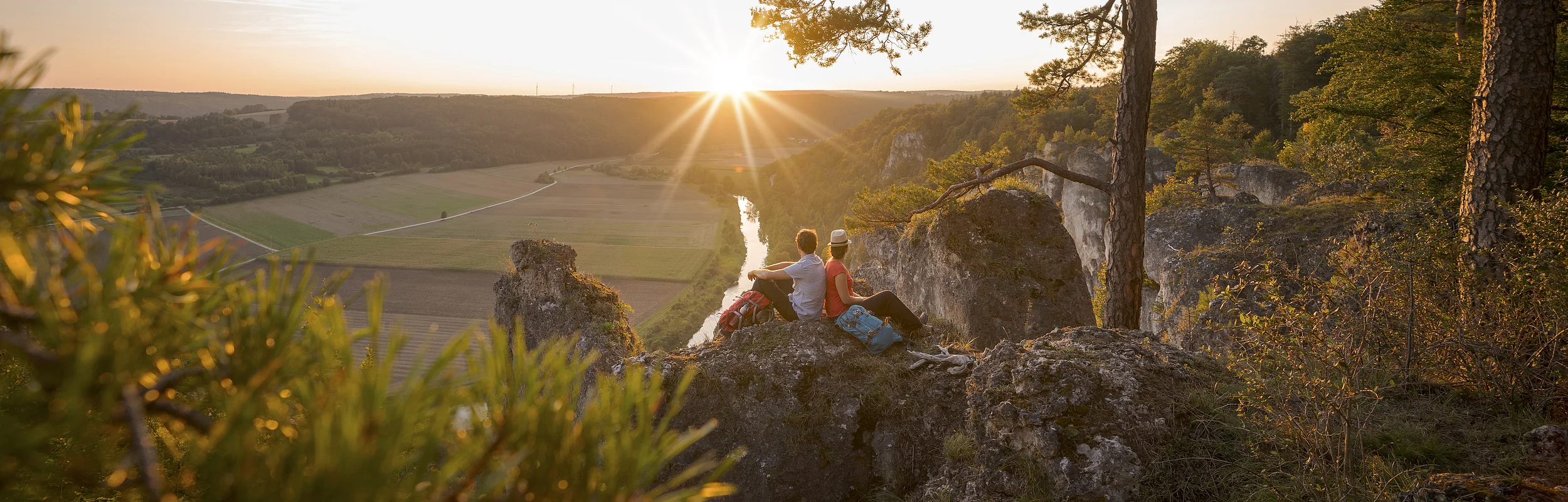 Zwei Personen sitzen auf Felsen und schauen bei Sonnenuntergang auf ein Flusstal mit Bäumen und Feldern.