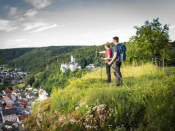Ein Frau und ein Mann beide in Wanderkleidung und mit Rucksäcken stehen auf dem grün bewachsenem Michelsberg und schauen auf Kipfenberg hinunter. Im Hintergrund ist die Burg Kipfenberg inmitten von grünem Wald etwas zu erkennen. Der Himmel ist leicht grau und bewölkt.