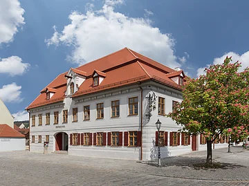 Weißes zweistöckiges Gebäude mit roten Fensterläden und Dach, blühender Baum rechts, gepflasterter Platz davor