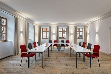 Konferenzraum mit U-förmiger Tischanordnung, roten und grauen Stühlen und Fenstern mit Vorhängen.