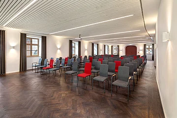 Konferenzraum mit Reihen von grauen und roten Stühlen, weißen Wänden und Holzboden.