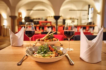 Tisch mit zwei Tellern Essen, zwei Gläsern Rotwein und gefalteten weißen Servietten in einem Restaurant mit Bogenfenstern.