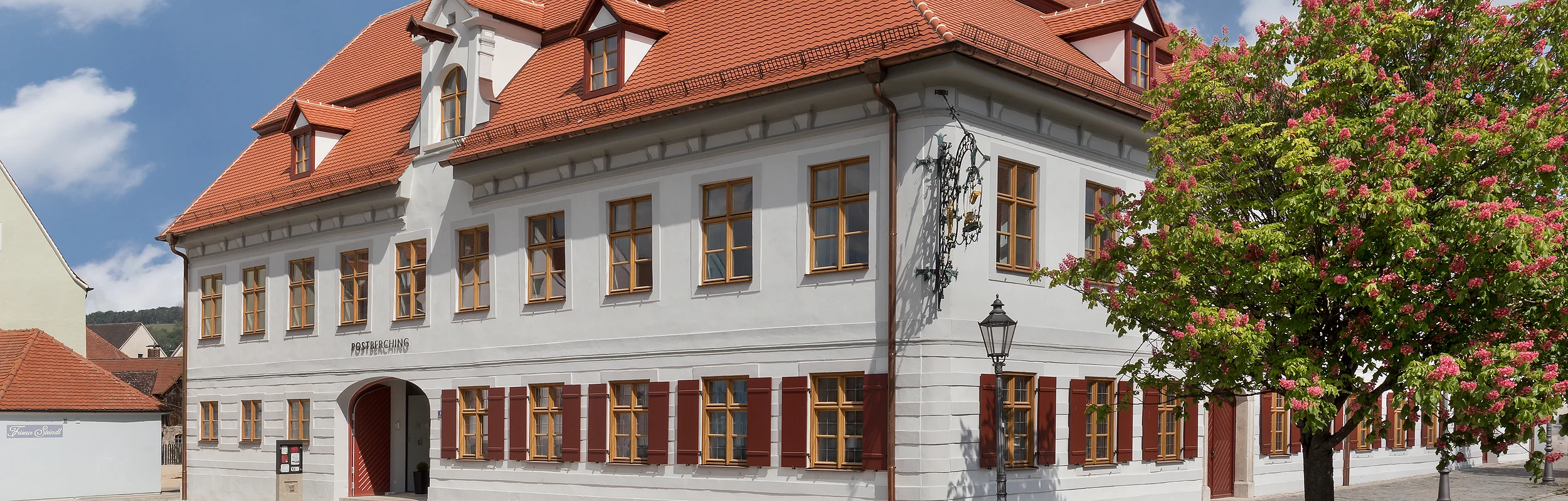 Weißes zweistöckiges Gebäude mit roten Fensterläden und Dach, blühender Baum rechts, gepflasterter Platz davor