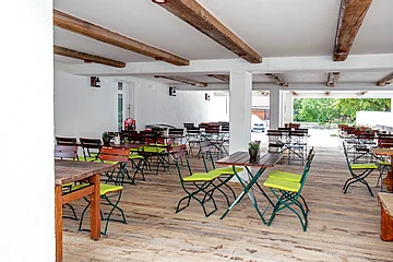 Überdachter Außenbereich mit Holzboden, Holztischen und Stühlen mit grünen Sitzkissen, weiße Wände und Decke mit Holzbalken.