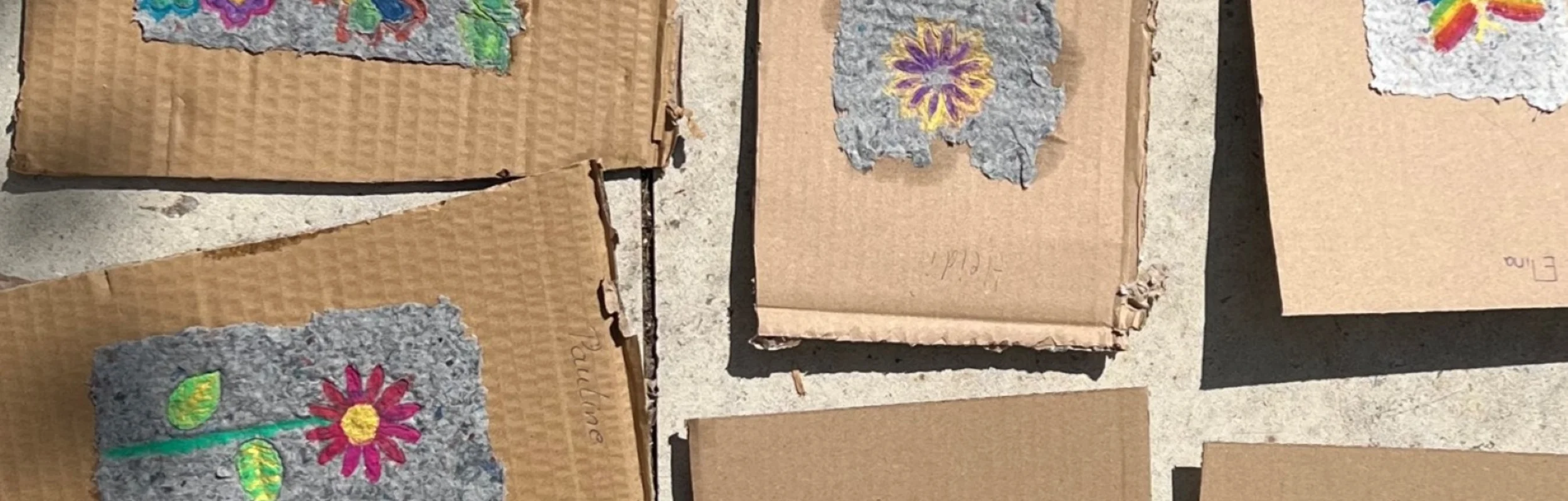 Mehrere Hand geschöpfte Papierstücke mit Blumen- und Schmetterlingsmotiven liegen auf braunen Kartonstücken auf einem Steinboden.