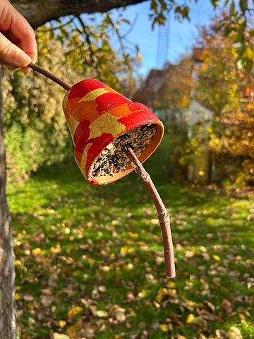 Hand hält bemalten Blumentopf mit Vogelfutter an einem Ast vor unscharfem Gartenhintergrund im Herbst