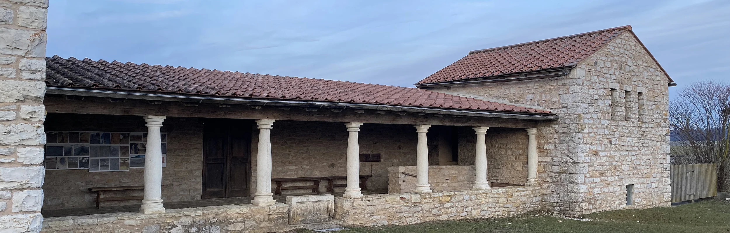 Steinernes Gebäude mit Säulen und rotem Ziegeldach vor bewölktem Himmel und Rasenfläche