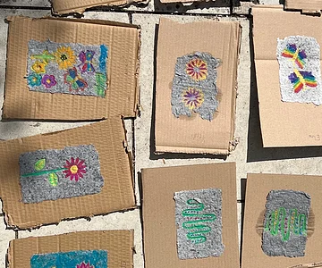 Mehrere Hand geschöpfte Papierstücke mit Blumen- und Schmetterlingsmotiven liegen auf braunen Kartonstücken auf einem Steinboden.