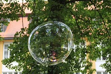 Seifenblasen Eine riesige Seifenblase schwebt in der Höhe, hinter ihr die grünen Äste eines Baumes. Durch diese ist ein Gebäude zu erkennen.