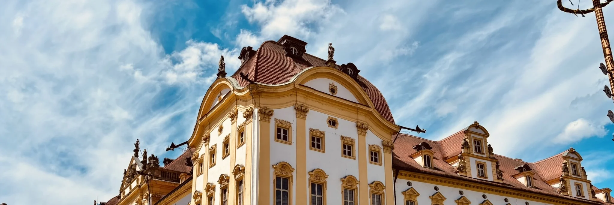 Zentrales Element des Bildes ist die Schlossresidenz Ellingen mit kräftigen Farben vor einem blauen Himmel. Links ist ein Pfeiler mit einer Statue zu sehen