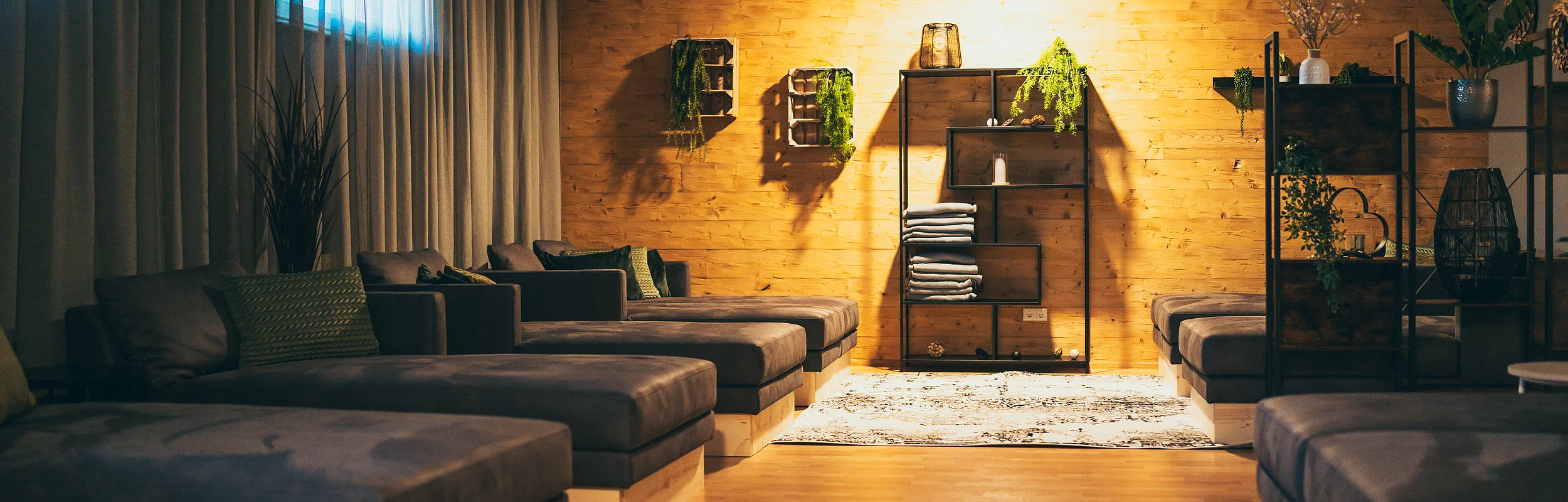 Raum mit mehreren gepolsterten Liegen, Holzwand, Regalen mit Pflanzen und Handtüchern, Vorhängen am Fenster.