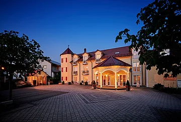 Beleuchtetes Hotelgebäude mit gepflastertem Vorplatz und Bäumen bei Abenddämmerung.