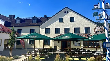 Außenansicht eines Gasthauses mit grünen Sonnenschirmen und Sitzgelegenheiten bei klarem Himmel.