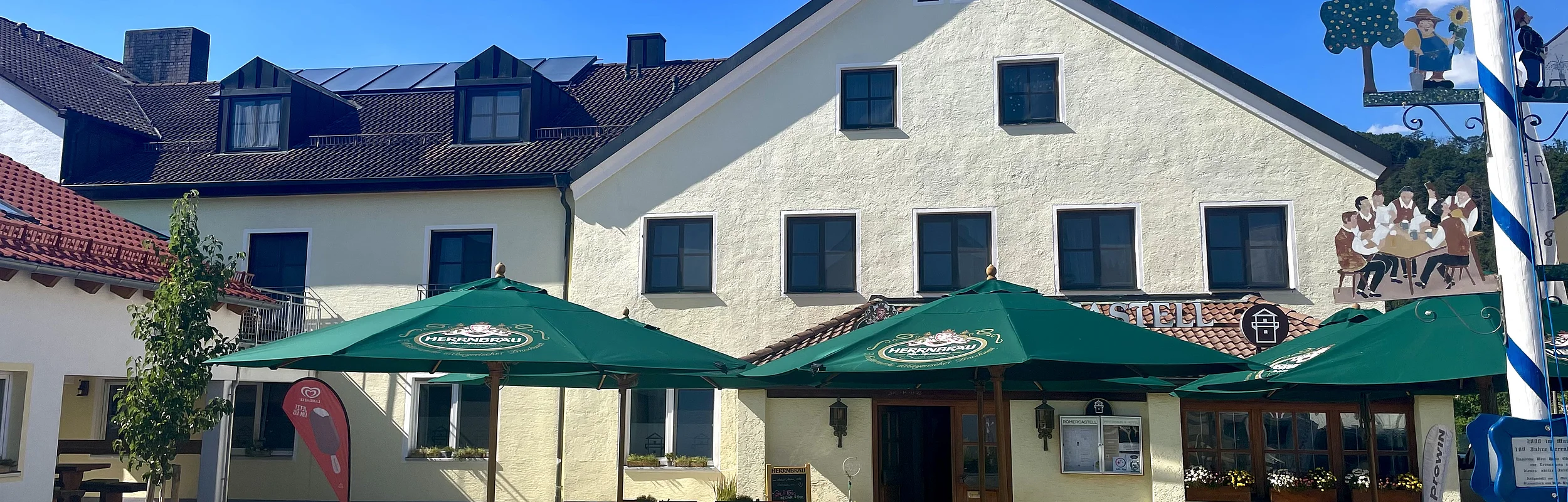 Außenansicht eines Gasthauses mit grünen Sonnenschirmen und Sitzgelegenheiten bei klarem Himmel.