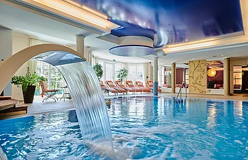 Innenaufnahme eines Schwimmbads mit Wasserfall, Liegestühlen und blauem Deckengewölbe mit Beleuchtung.