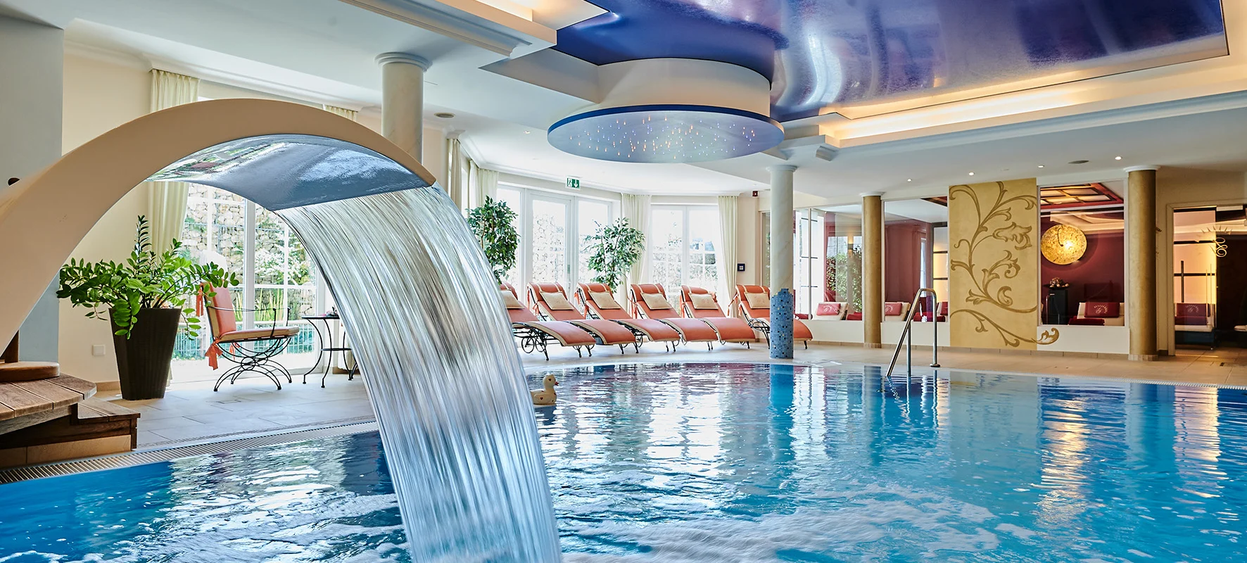 Innenaufnahme eines Schwimmbads mit Wasserfall, Liegestühlen und blauem Deckengewölbe mit Beleuchtung.