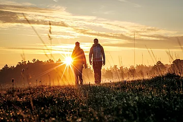 Zwei Personen gehen bei Sonnenaufgang über eine Wiese mit hohem Gras und Bäumen im Hintergrund.