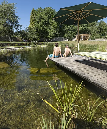 Zwei Jungen sitzen auf einem Holzsteg am Ufer eines Naturteichs unter blauem Himmel mit grünem Sonnenschirm.
