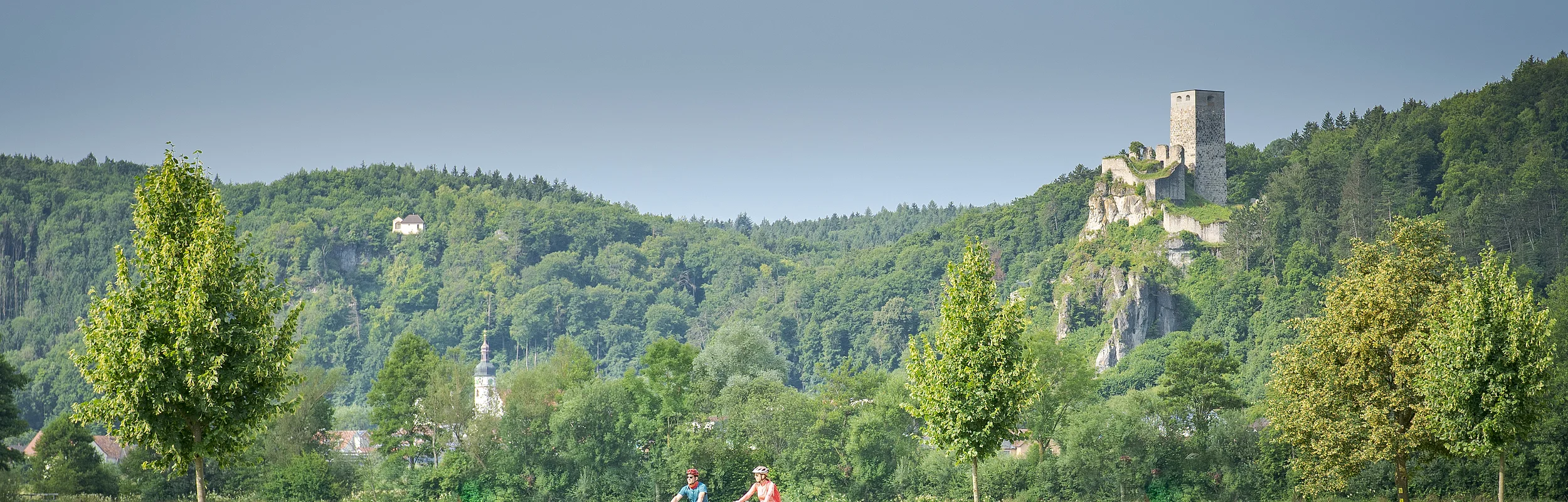 Zwei Radfahrer auf einem Weg vor grüner Wiese, Bäumen und bewaldetem Hügel mit Burgruine im Hintergrund.