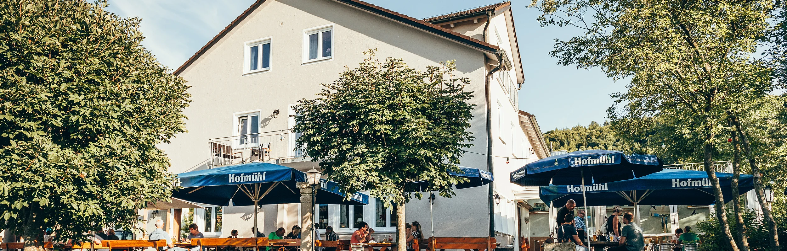Gastgarten mit blauen Sonnenschirmen und Holzbänken vor einem weißen Gebäude an einem sonnigen Tag