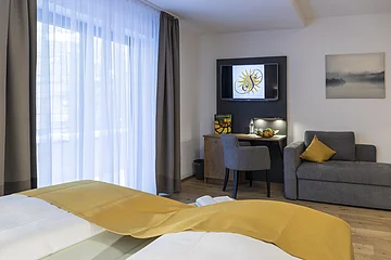 Hotelzimmer mit Bett, Schreibtisch, Fernseher, grauem Sessel und Sofa mit gelbem Kissen, helle Vorhänge am Fenster.