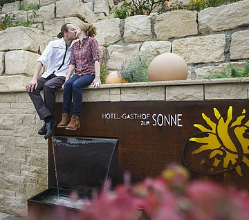 Paar sitzt auf Mauer über Brunnen mit Schriftzug „Hotel-Gasthof zur Sonne“ und küsst sich.
