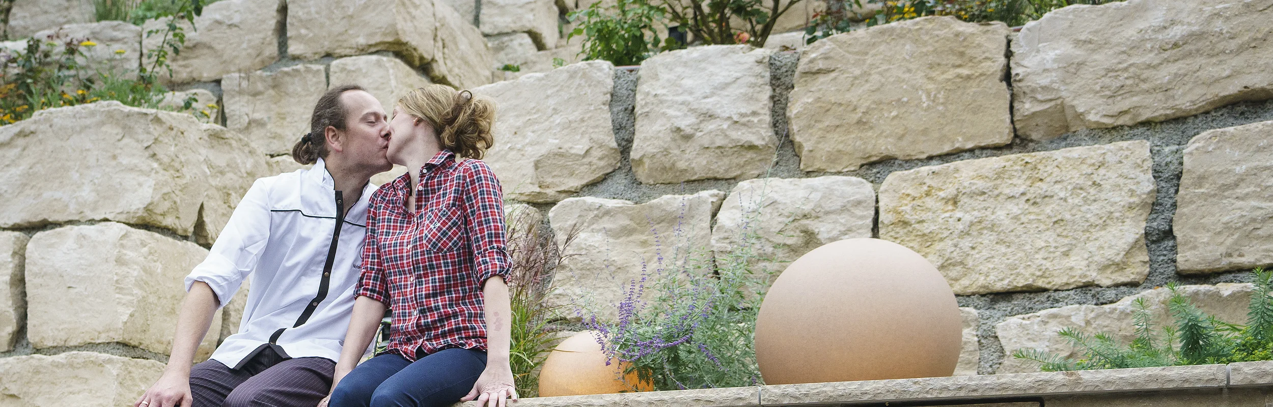 Paar sitzt auf Mauer über Brunnen mit Schriftzug „Hotel-Gasthof zur Sonne“ und küsst sich.
