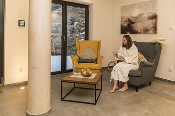 Frau im weißen Bademantel sitzt auf grauem Sessel, hält Glas, neben gelbem Sessel und Holztisch mit Obstschale.