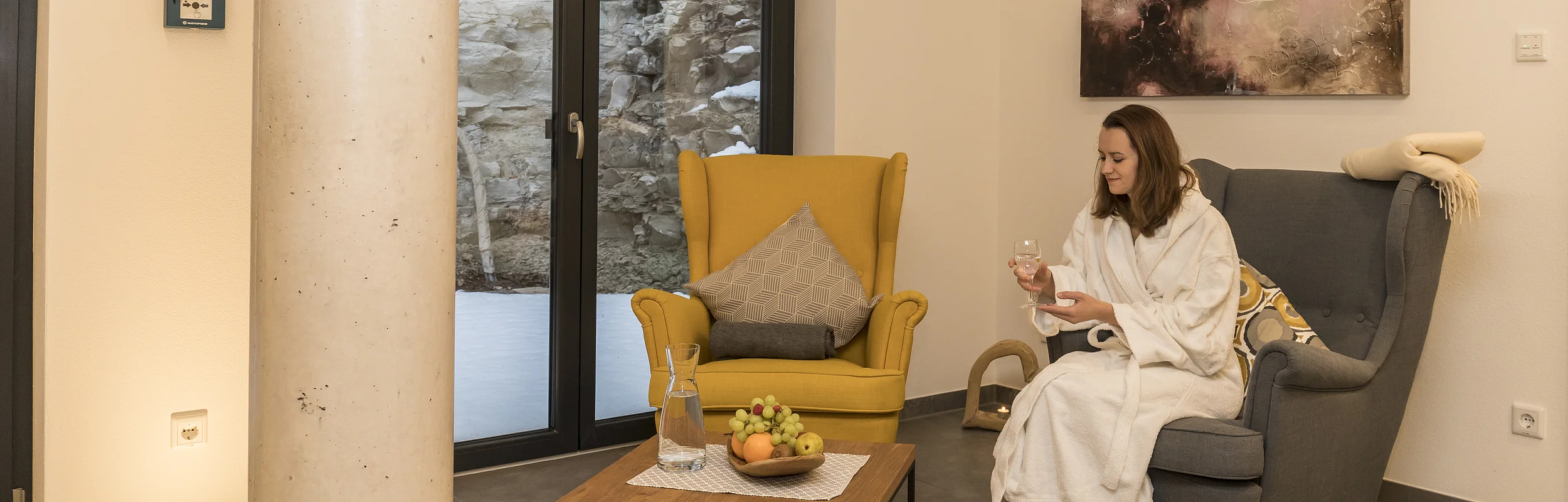 Frau im weißen Bademantel sitzt auf grauem Sessel, hält Glas, neben gelbem Sessel und Holztisch mit Obstschale.