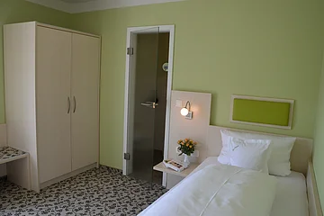 Hotelzimmer mit Einzelbett, grün gestrichener Wand, Schrank, Glastür und Nachttisch mit Lampe und Blumen.