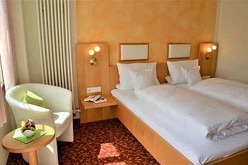 Hotelzimmer mit zwei Einzelbetten, Nachttisch, Sessel, kleinem Tisch und Obstschale bei Tageslicht.