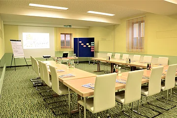 Tagungsraum mit U-förmiger Tischanordnung, weißen Stühlen, Flipchart und Projektor mit Leinwand.