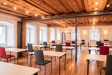 Seminarraum mit Holzboden, Holzbalkendecke, weißen Tischen und roten sowie grauen Stühlen, Fenster mit weißen Vorhängen.