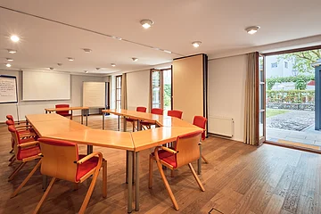 Konferenzraum mit U-förmigen Holztischen, roten Stühlen, Flipcharts und offenem Ausgang zum Außenbereich.