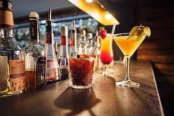 Bar mit verschiedenen Spirituosenflaschen und drei Cocktails auf der Theke, darunter ein brauner Drink mit Zimtstange.