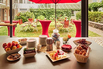 Buffet mit Obst, Gebäck, Nüssen und Tellern auf Tisch vor Garten mit roten Stehtischen und Stühlen